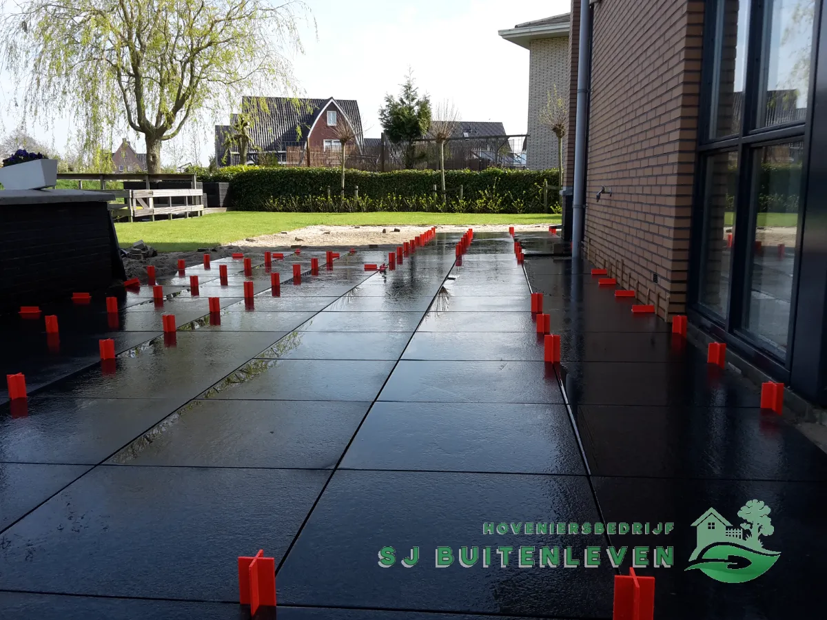 Aanleg van grootformaat terrastegels in donkere kleur tegen de gevel in Heerhugowaard, met tegelkruisjes voor een strakke voegverdeling tijdens het leggen.